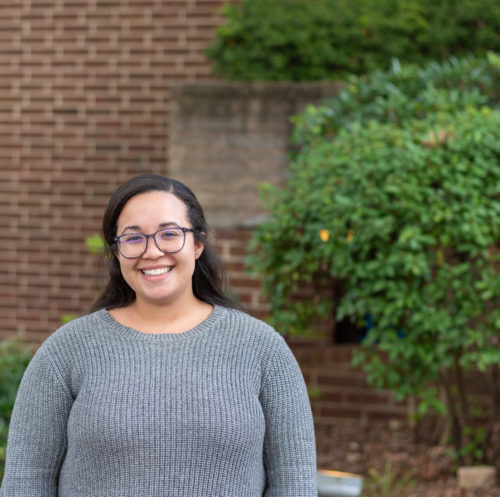 Head shot of Kristina Smith: mixed race woman wearing glasses and a sweater standing in front of a brick wall and a tree
