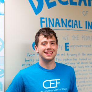 Zach Meredith headshot: white man wearing bright blue CEF t-shirt standing in front of a white wall with bright blue text