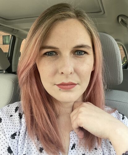 A white woman with below shoulder length straight hair, dyed pink at the bottom sits in a car wearing a polka dot white and black shirt. She is not smiling or frowning and staring directly into the camera. One arm is visible. Her hand is making a loose fist against her collarbone.